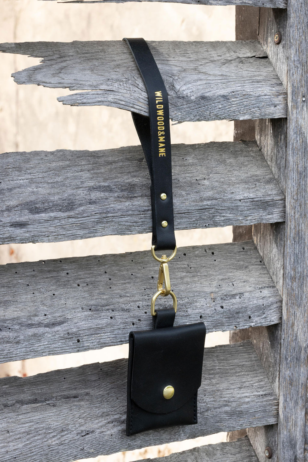 Black wallet with wrist strap and brass hardware on a wooden shutters. 3/4 view