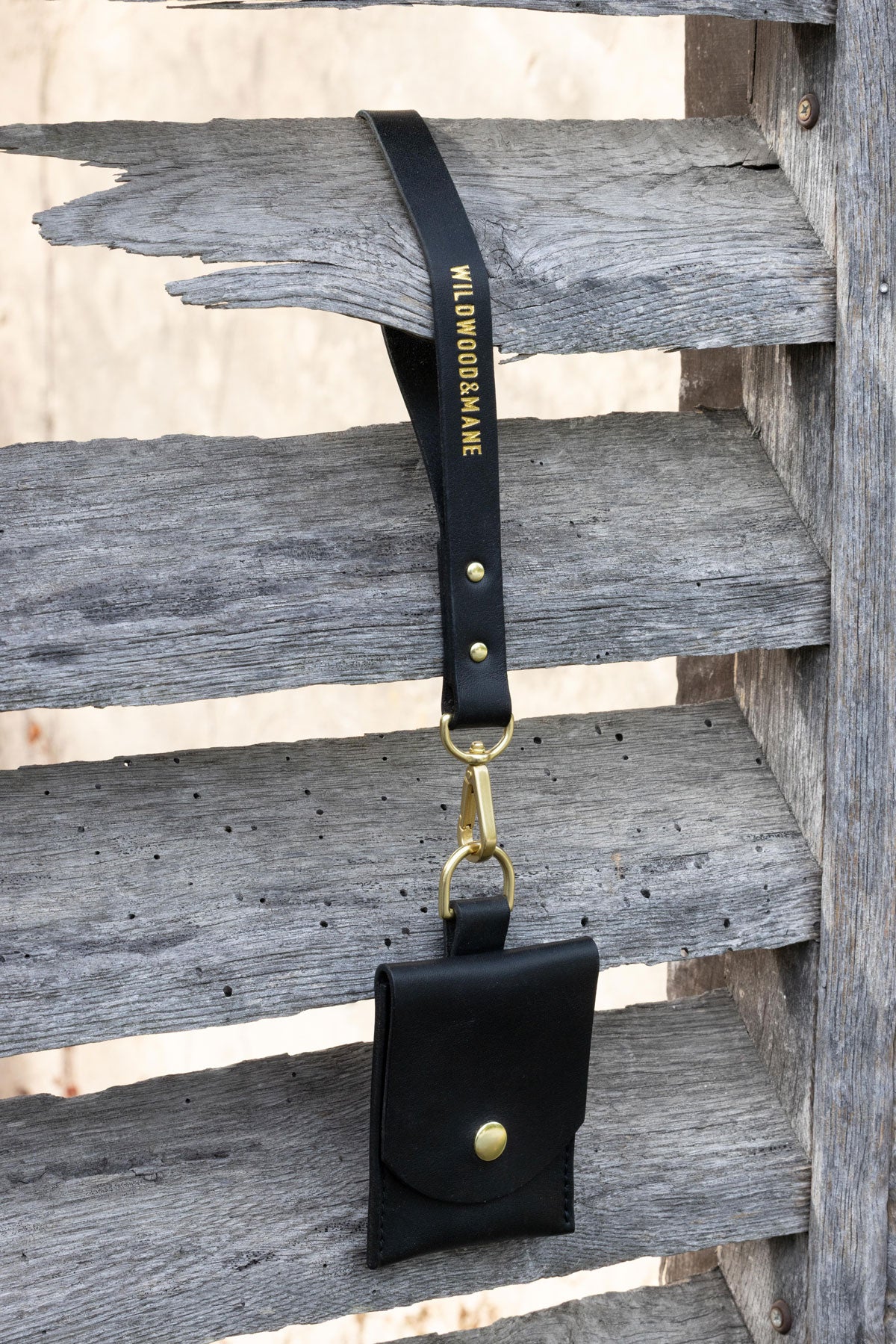 Black wallet with wrist strap and brass hardware on a wooden shutters. 3/4 view