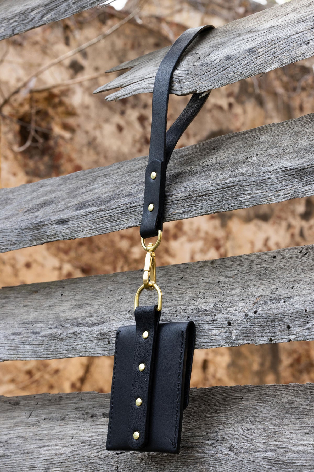 Black leather wallet with wrist strap and brass hardware on a wooden shutters. Back 3/4 view