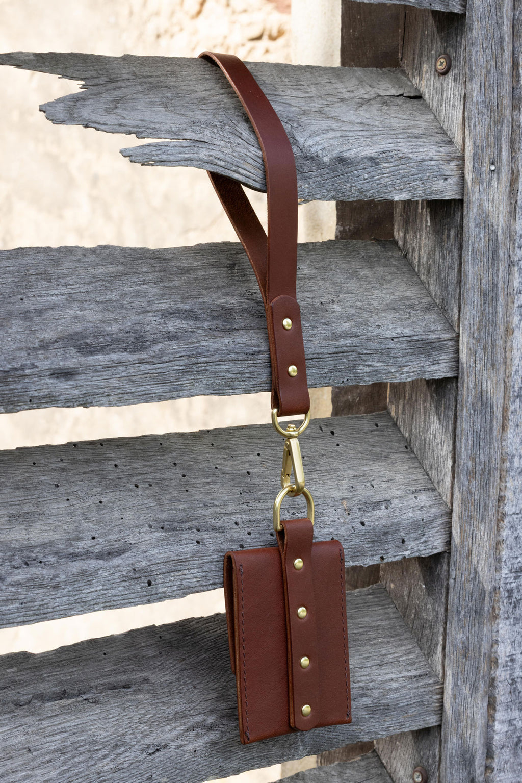 Brown leather wallet with wrist strap with gold hardware on a wooden shutters. Back view