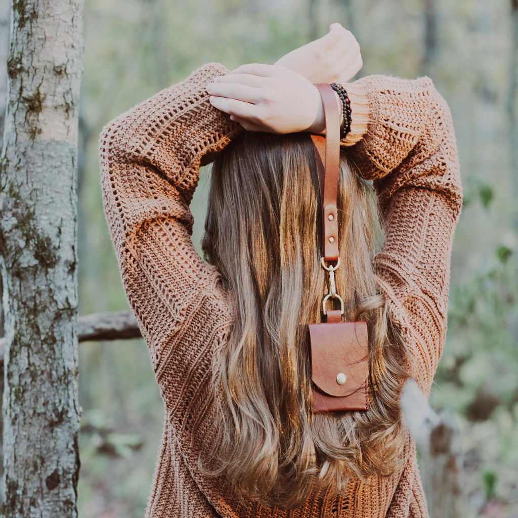 Person wearing a brown knitted sweater holding a brown leather wallet with wrist strap in a forest setting